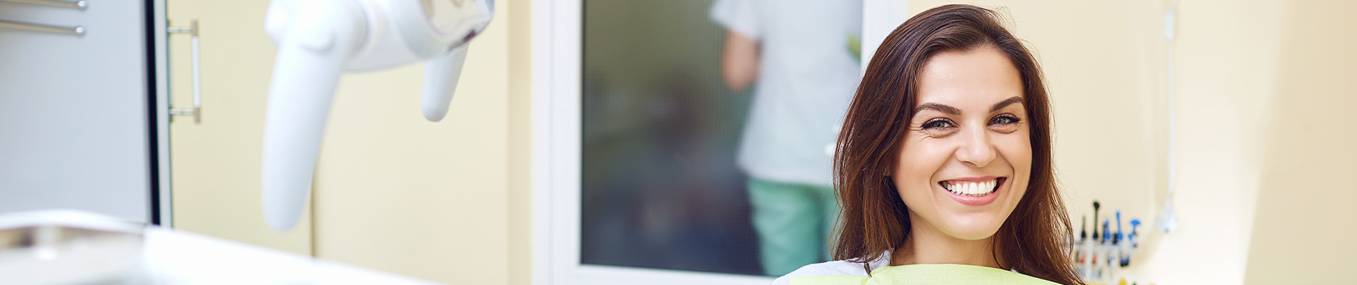 A person is seated in a dental chair, receiving care from a dental professional who stands behind them.